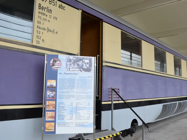 ALSTOM Werksmuseum_HD_16zu9.jpg Lila-cremefarbener historischer Zugwaggon mit geöffneter Tür und einer Informationstafel.