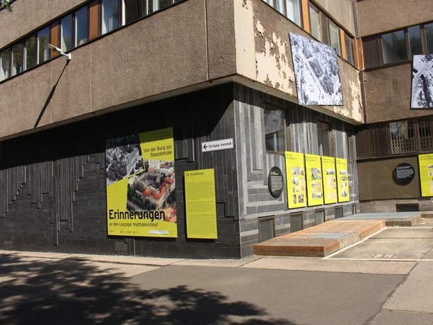 Wystawa plenerowa z wielkoformatowymi tablicami informacyjnymi i historycznymi zdjęciami zamontowanymi wzdłuż fasady budynku, żywo prezentująca historię Matthäikirchhof.