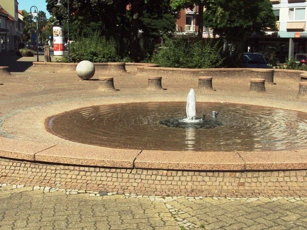 Brunnen_Bohlweg_SZ-Bad_HD_16zu9.JPG Ein runder Brunnen mit einer kleinen Fontäne auf einem gepflasterten Platz, umgeben von Bäumen.