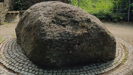 Brunnen_Irenenquelle_SZ-Bad_HD_16zu9.jpg Ein großer, runder Findling ruht auf einem gepflasterten Platz, umgeben von einem Gitterzaun.