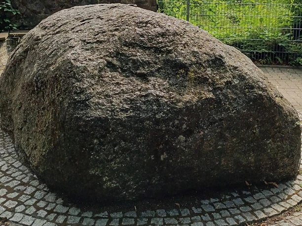 Ein großer, runder Findling ruht auf einem gepflasterten Platz, umgeben von einem Gitterzaun.