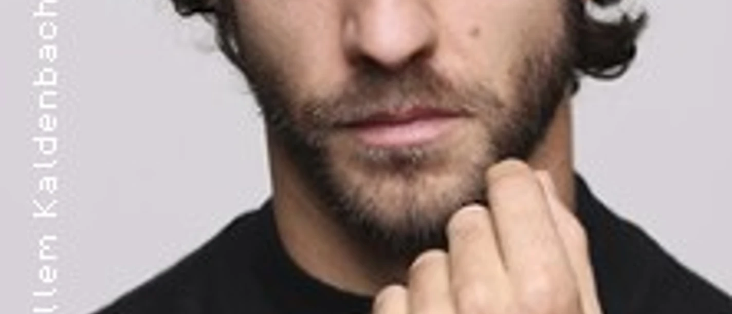 Porträt eines Mannes mit leichtem Bart, direktem Blick und erhobener Hand vor hellem Hintergrund
