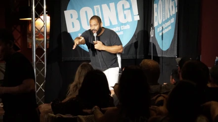 BOING! English Comedy Night Comedian on stage with microphone in front of audience, in front of blue BOING! banner in bright spotlight.