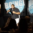 BOING! English Comedy Night Comedian auf Bühne mit Mikrofon vor Publikum, vor blauem BOING!-Banner in hellem Scheinwerferlicht.
