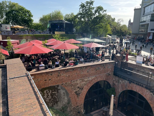 View from above of the Moritzbastei with its outdoor seating area, where numerous guests can be seen enjoying the sun, with an open-air stage in the background.