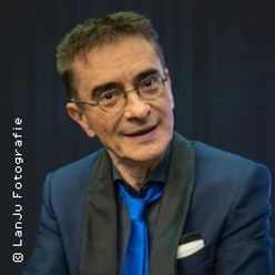 Ein älterer Mann im blauen Anzug mit Brille vor dunklem Hintergrund