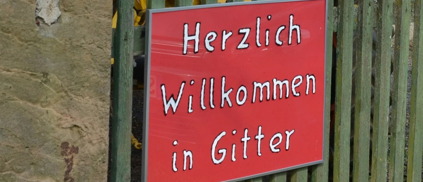 Willkommen in Gitter_HD_16zu9.JPG Rotes Schild am grünen Holzzaun mit weißer Schrift: Herzlich Willkommen in Gitter.