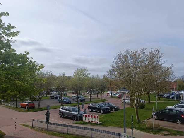 Öffentlicher Parkplatz mit geparkten Autos, umgeben von Bäumen und Rasen unter Wolkenhimmel.