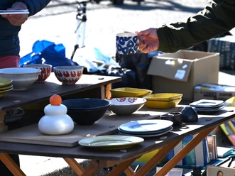 Flohmarkt.png Person hält eine blaue Tasse an einem Flohmarktstand mit bunten Keramiktellern und Schalen.