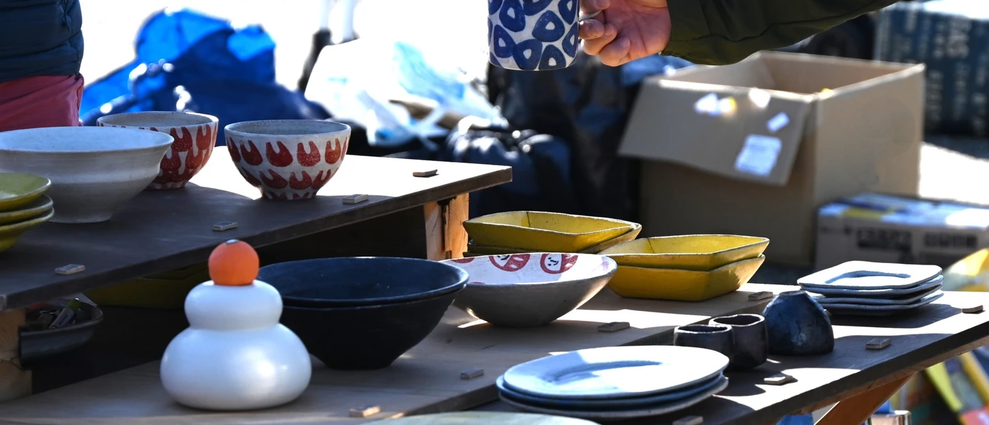 Flohmarkt.png Person hält eine blaue Tasse an einem Flohmarktstand mit bunten Keramiktellern und Schalen.