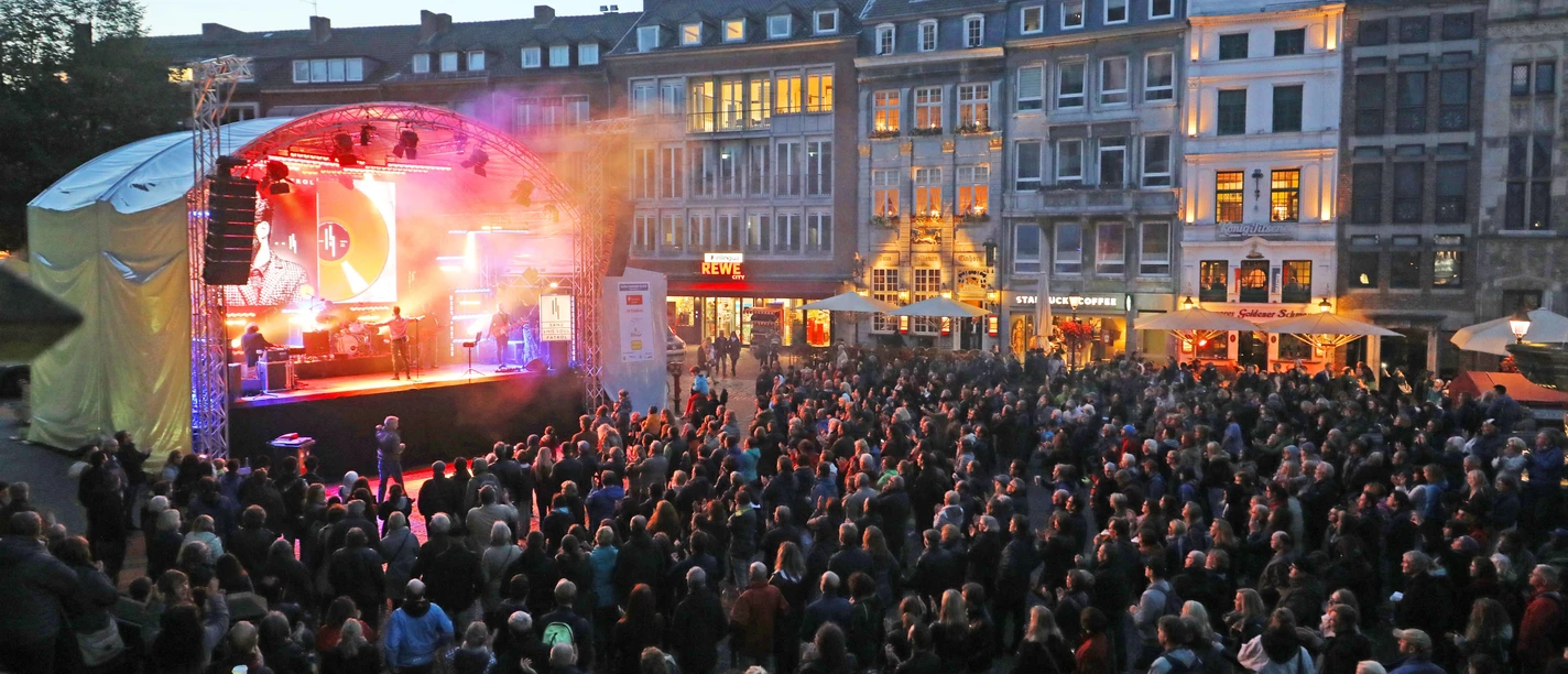 Auf dem Marktplatz ist eine Bühne aufgebaut, vor der viele Zuschauer*innen versammelt sind und einem Konzert lauschen.