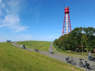 Deich beim Campener Leuchtturm Radfahrer auf einem grünen Deich vor dem rot-weißen Campener Leuchtturm bei blauem Himmel.