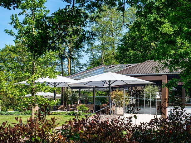 Clubhaus mit Terrasse und Sonnenschirmen am Golfplatz, umgeben von Bäumen und Rasen
