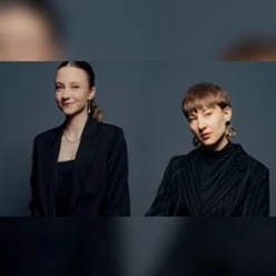 Zwei Personen in dunkler Kleidung vor dunklem Hintergrund, frontal und halbseitig dargestellt.
