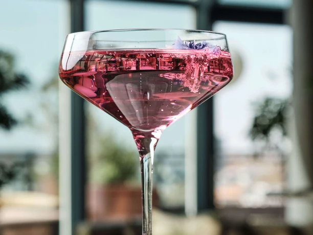 Bar Botanik - Signature Drink Rosa Cocktail in einer flachen Glaskelchform vor großen Fenstern mit weich verschwommenem Hintergrund.