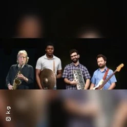Vier Musiker mit Saxophon, Becken, Keyboard und Gitarre vor dunklem Hintergrund.