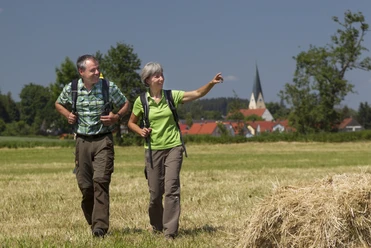 Wanderer bei Pfakofen Zwei Wandernde auf einer Wiese, im Hintergrund ein Dorf mit Kirchturm unter blauem Himmel.