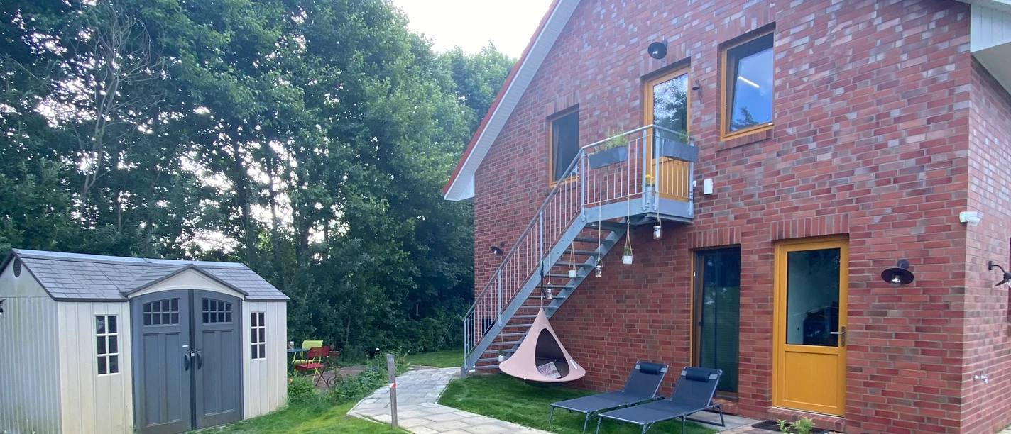 Eingang zum Worpsweder Glück Ferienwohnung im roten Backsteinhaus mit Außentreppe, Garten, Liegestühlen und kleiner Hütte.