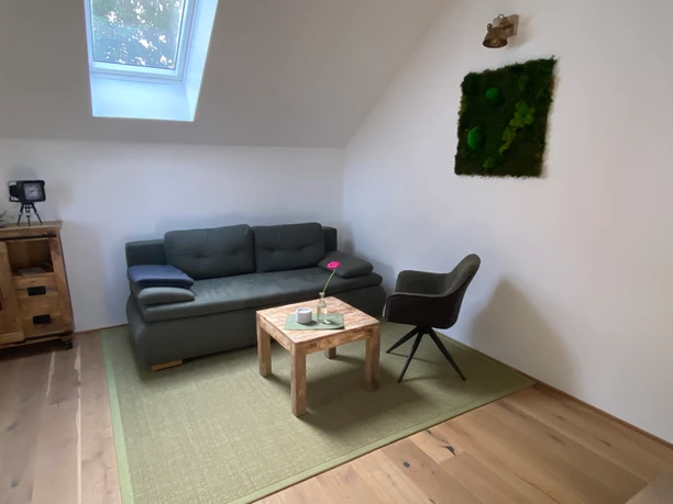 Im Wohnzimmer Ein helles Wohnzimmer in einer Ferienwohnung mit Sofa, Sessel, Holztisch und einem Dachfenster.