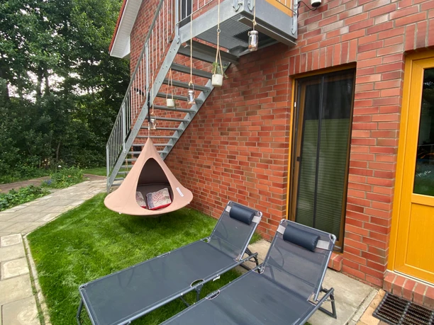 Auszeit im Garten Backsteinhaus mit Außentreppe, darunter ein Hängesessel und zwei Sonnenliegen auf dem Rasen.