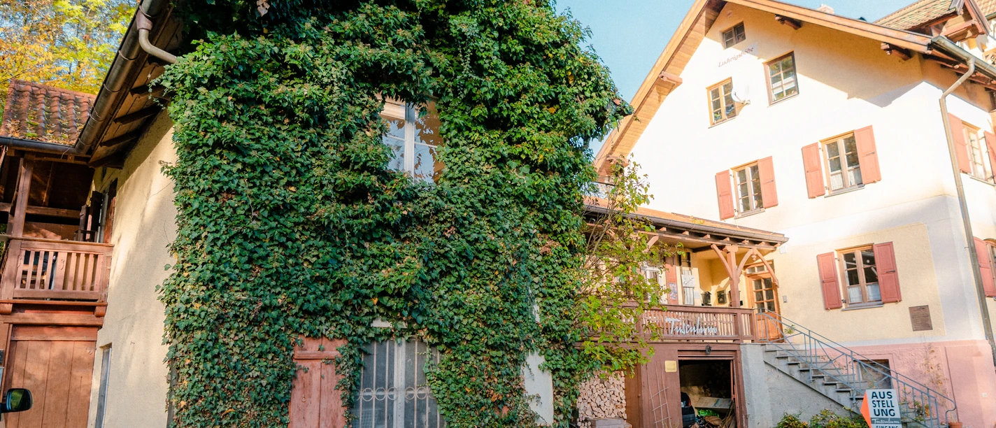 Tusculum @Tourist Information Murnau.jpg Das Bild zeigt das Christoph-Probst-Geburtshaus in Murnau.. Ein kleines Haus mit Efeu neben einem größeren Gebäude an einem herbstlichen Hof.
