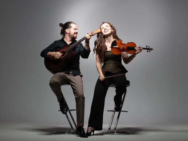 Festival aufm Platz Duo Lea Brückner und Gábor Ladányi Ein fröhliches Musiker-Duo auf Barhockern: Er spielt Gitarre, sie hält lächelnd eine Geige.
