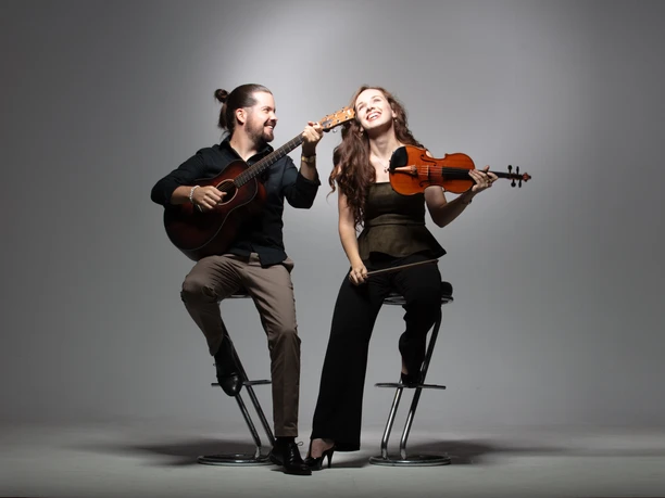 Brueckner_Ladanyi-1.jpg Ein fröhliches Musiker-Duo auf Barhockern: Er spielt Gitarre, sie hält lächelnd eine Geige.
