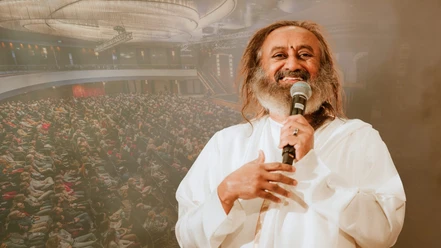 Gurudev Sri Sri Ravi Shankar Ein sprechender Mann in weißer Kleidung vor großem Publikum in einem Saal.