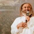 Gurudev Sri Sri Ravi Shankar A speaking man in white clothes in front of a large audience in a hall.