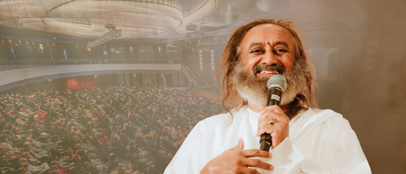 Gurudev Sri Sri Ravi Shankar A speaking man in white clothes in front of a large audience in a hall.
