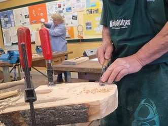 Freier Schnitzkurs Hände eines Mannes mit einem Holzmeißel, der an einem Holzstück arbeitet, im Hintergrund eine Person.
