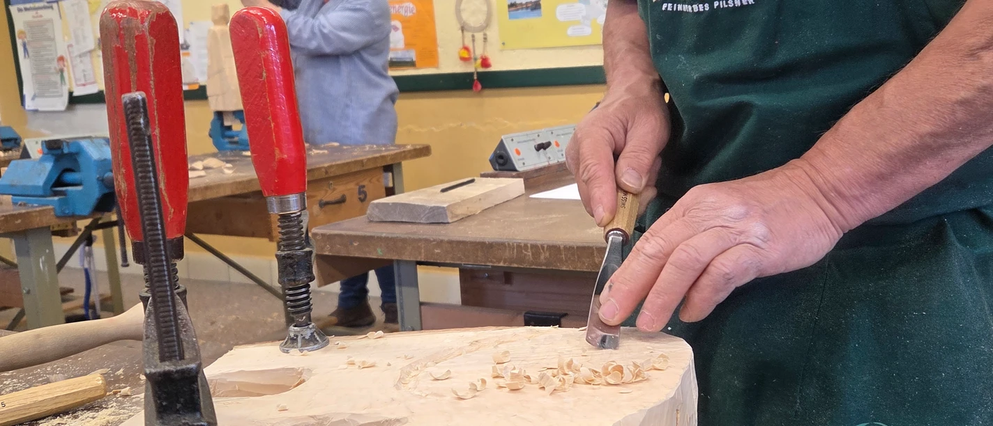 Freier Schnitzkurs Hände eines Mannes mit einem Holzmeißel, der an einem Holzstück arbeitet, im Hintergrund eine Person.