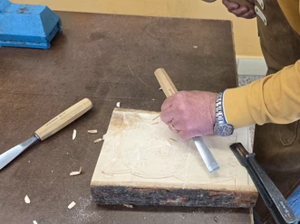 Schnitzkurs in Schnitzbude Ein Handwerker schnitzt mit einem Meißel in ein Holzstück auf einem Tisch.
