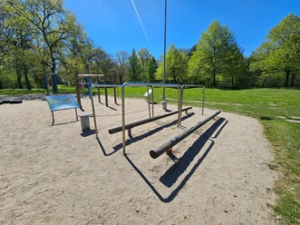 Gesundheitspark Speckenbüttel 20260429_135523 (c) Tanja Mehl_Erlebnis Bremerhaven_WEB.jpg Fitnessgeräte mit Barren und Balancierbalken auf Sand im grünen Park unter blauem Himmel.
