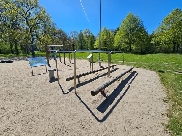 Gesundheitspark Speckenbüttel 20260429_135523 (c) Tanja Mehl_Erlebnis Bremerhaven_WEB.jpg Fitnessgeräte mit Barren und Balancierbalken auf Sand im grünen Park unter blauem Himmel.
