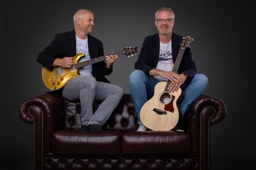 Double Tuff.jpg Zwei Musiker sitzen auf einem Ledersofa. Der eine hält einen Bass, der andere eine Gitarre in der Hand