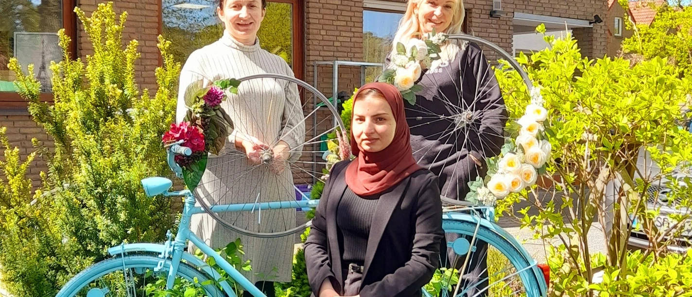 Radlertreff_SHS_Mai2026.jpg Drei Frauen vor Backsteingebäude mit blauem Fahrrad und Blumendeko