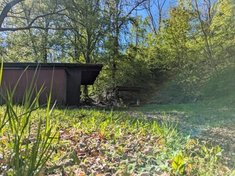 Schutzhütte Bartshausen, Panoramaweg Dunkle hölzerne Schutzhütte mit Rastplatz inmitten von grünen Bäumen bei strahlendem Sonnenschein.