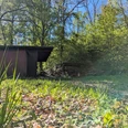 Schutzhütte Bartshausen, Panoramaweg Dunkle hölzerne Schutzhütte mit Rastplatz inmitten von grünen Bäumen bei strahlendem Sonnenschein.