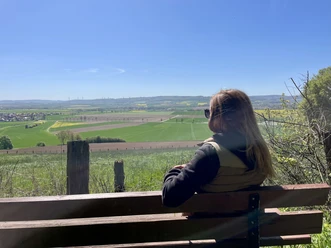 Rastplatz, Ausblick, Ahlsburg, Einbeck, Panoramaweg Frau blickt von einer Holzbank über weite, sonnige Felder in das grüne Leinetal.