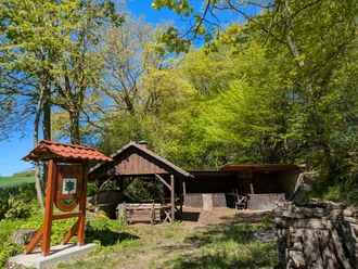 Grillhüte Avendshausen, Panoramaweg, CC0 (3).jpg Holzschild Grillhütte Avendshausen vor einer Holzhütte mit Brennholz am sonnigen Waldrand.