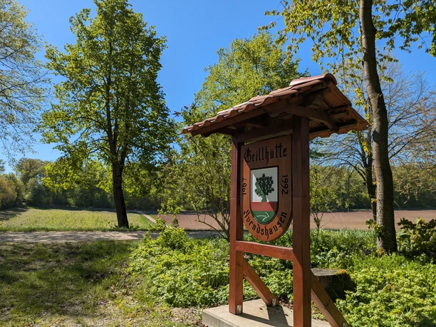 Grillhüte Avendshausen, Panoramaweg Ein überdachtes Holzschild mit Wappen zur Grillhütte Avendshausen an einem sonnigen Waldrand.