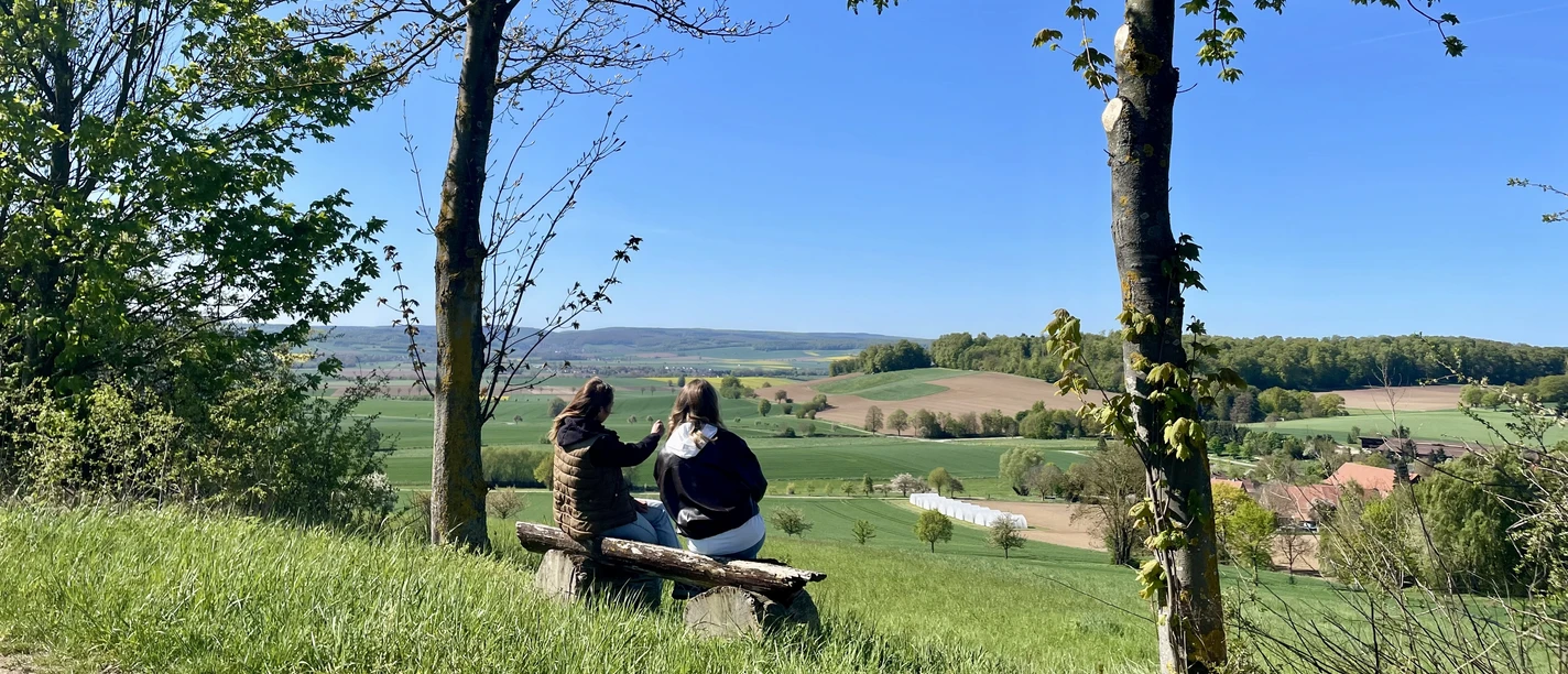To kvinder sidder på en træbænk og nyder panoramaudsigten over de vidtstrakte enge og marker.
