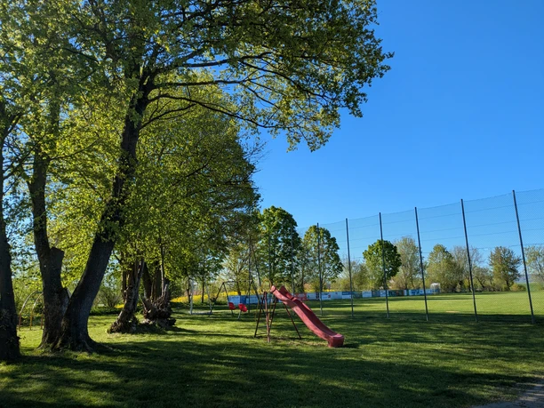 Sonniger Spielplatz mit roter Rutsche und Schaukel unter großen Bäumen am Sportplatz.