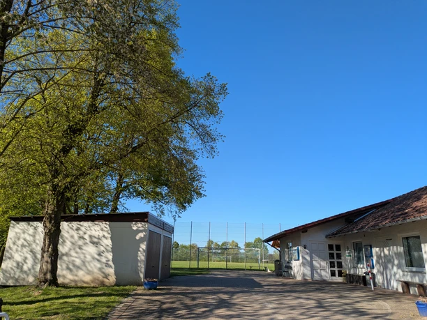 Sportplatz, Start, Vardeilsen, Panoramaweg Vorplatz am Sportheim mit großem Baum und Blick auf das Fußballfeld unter blauem Himmel.
