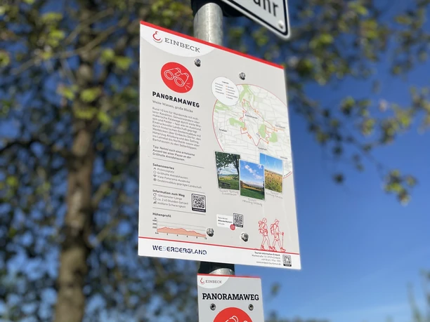 Starttafel, Parkplatz, Panoramaweg Weiße Informationstafel mit Karte und Wegweiser für den Panoramaweg vor blauem Himmel.
