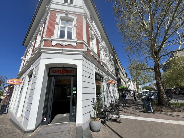 Eingang von Mam's Burger an Straßenecke mit Außensitzplätzen und Bäumen