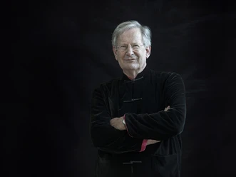 Sir John Eliot Gardiner Älterer Mann mit grauem Haar, verschränkten Armen und dunkler Kleidung vor schwarzem Hintergrund.