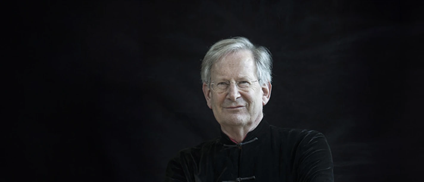 Sir John Eliot Gardiner Older man with gray hair, folded arms and dark clothes in front of a black background.