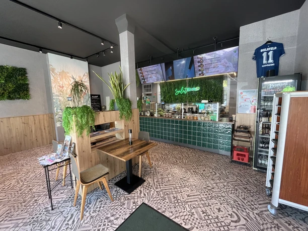 Innenraum einer modernen Salatbar mit Holztischen, grüner Theke und Pflanzenwand.
